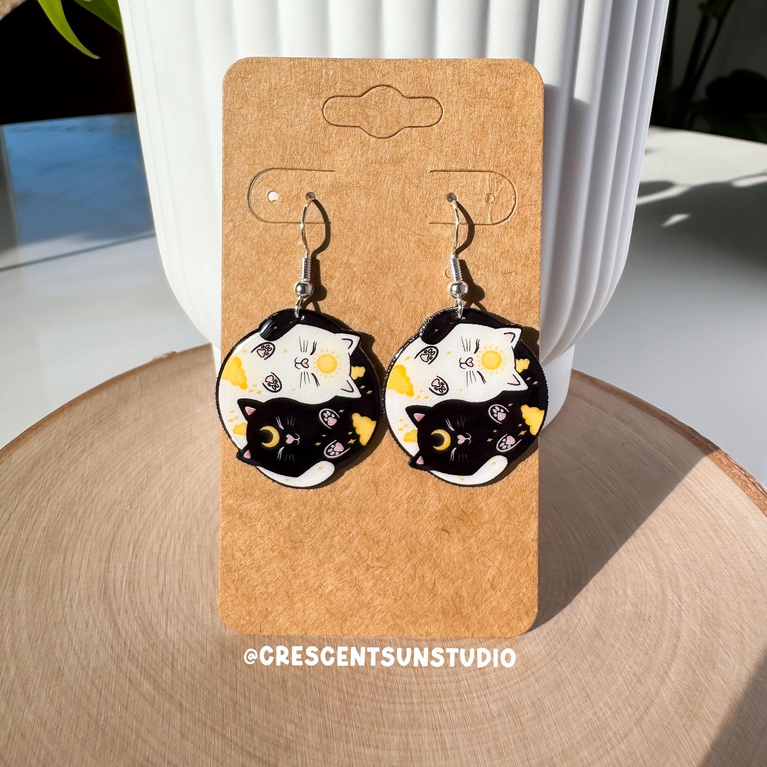 A pair of dangle earrings are displayed on a brown colored card. The hook for the earrings is silvertone. The earrings themselves are round and depict a black cat and a white cat curled up in a yin yang symbol. The black cat has a yellow crescent moon between her eyes and the white cat has a sun symbol between her eyes. Both smiling cats have two paws pointed outward, so you can see their pink toe pads.

In this image, the display card is propped up against a white plant pot.