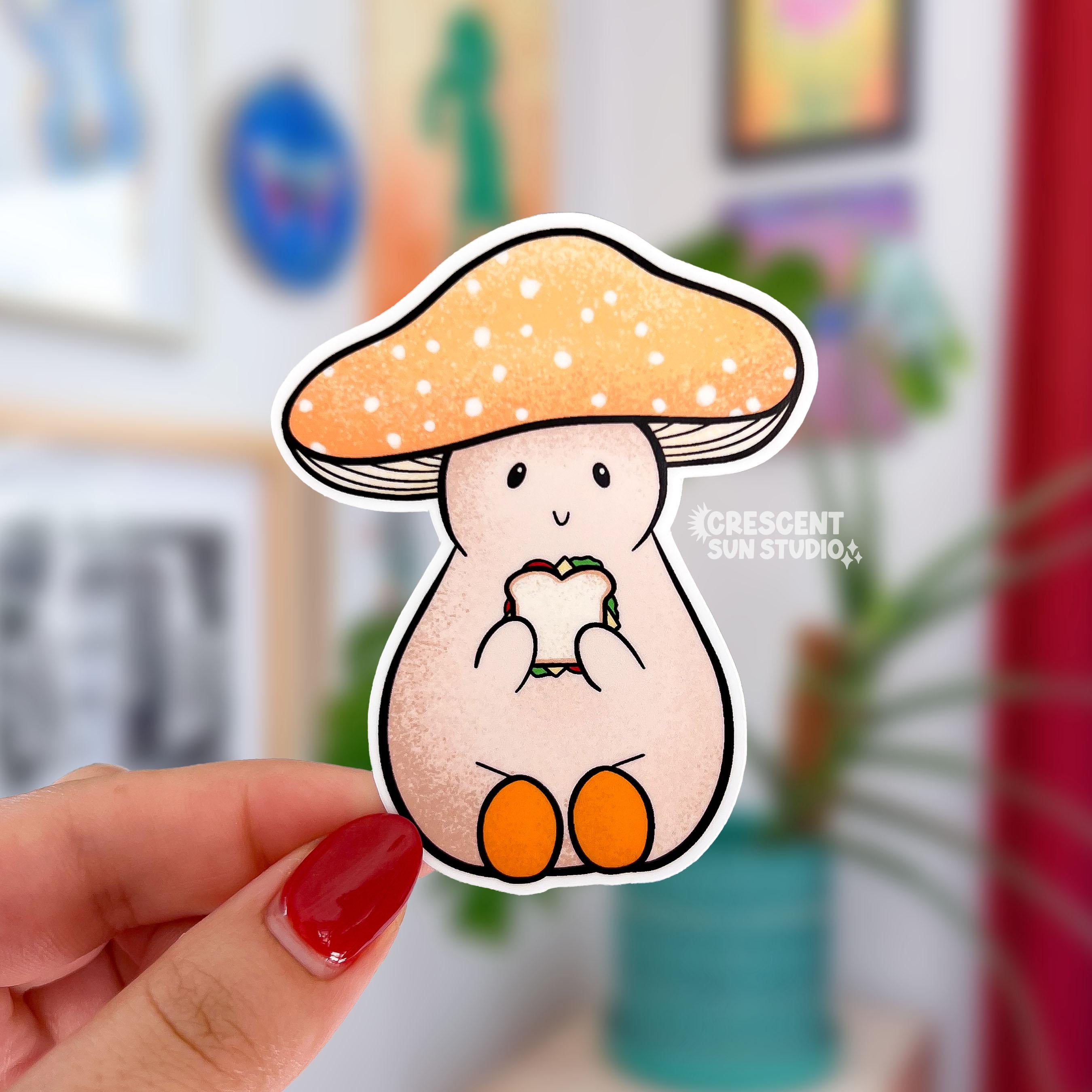 A die cut sticker of a pale mushroom character. His mushroom hate is a warm yellow with white spots. He is happily eating a sandwich. 