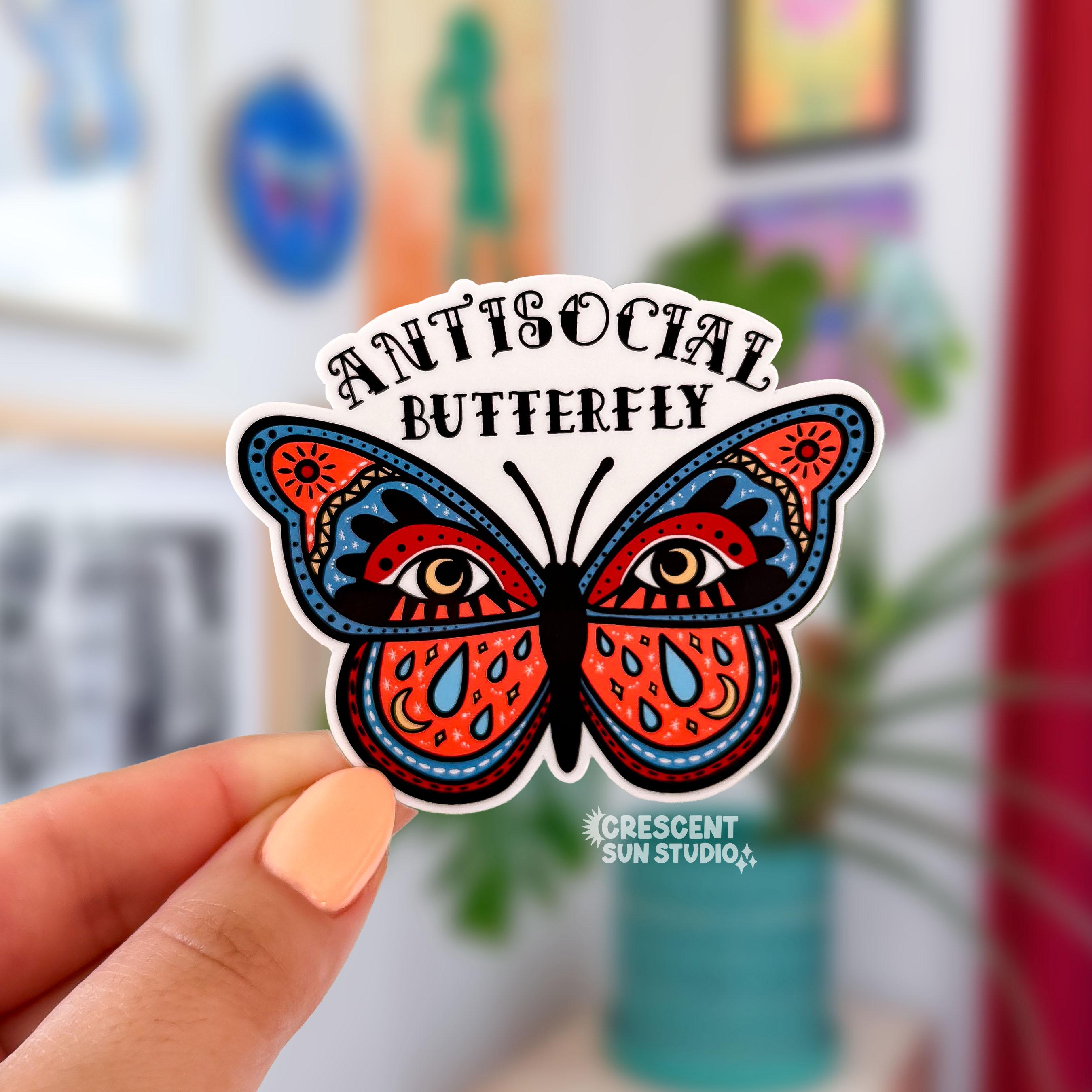 Die cut sticker of a boldly illustrated butterfly with large eyes on its wings and celestial details including moons, teardrops, and suns, with 'Antisocial Butterfly' arched above in decorative lettering.