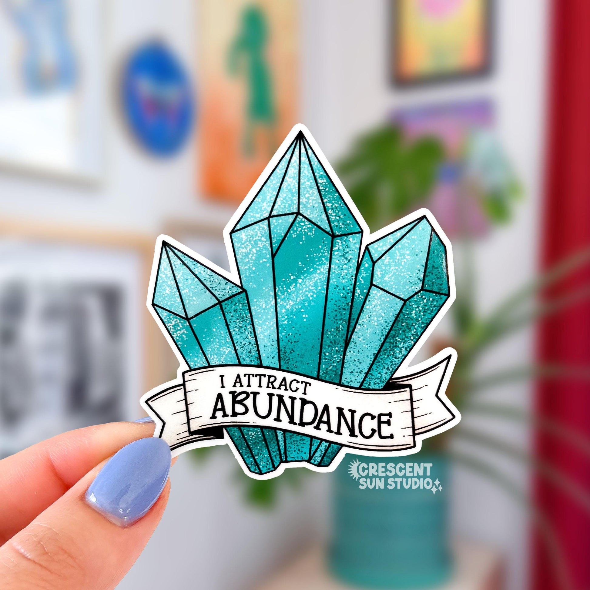 A sticker in the shape of three turquoise shaded crystals. At the bottom is a white banner with black text that reads "I attract abundance"