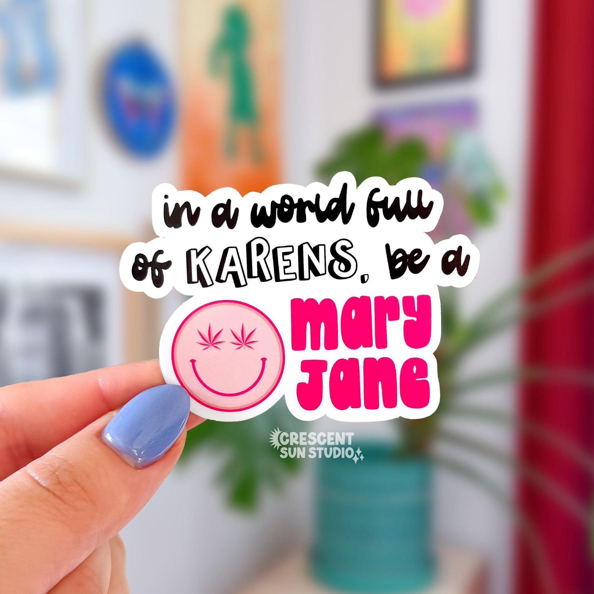 Die cut sticker with black text that reads, "in a world full of Karens, be a" and then there is a pink smiley face with cannabis leaves for eyes, and next to it in bold pink text it reads "mary jane."