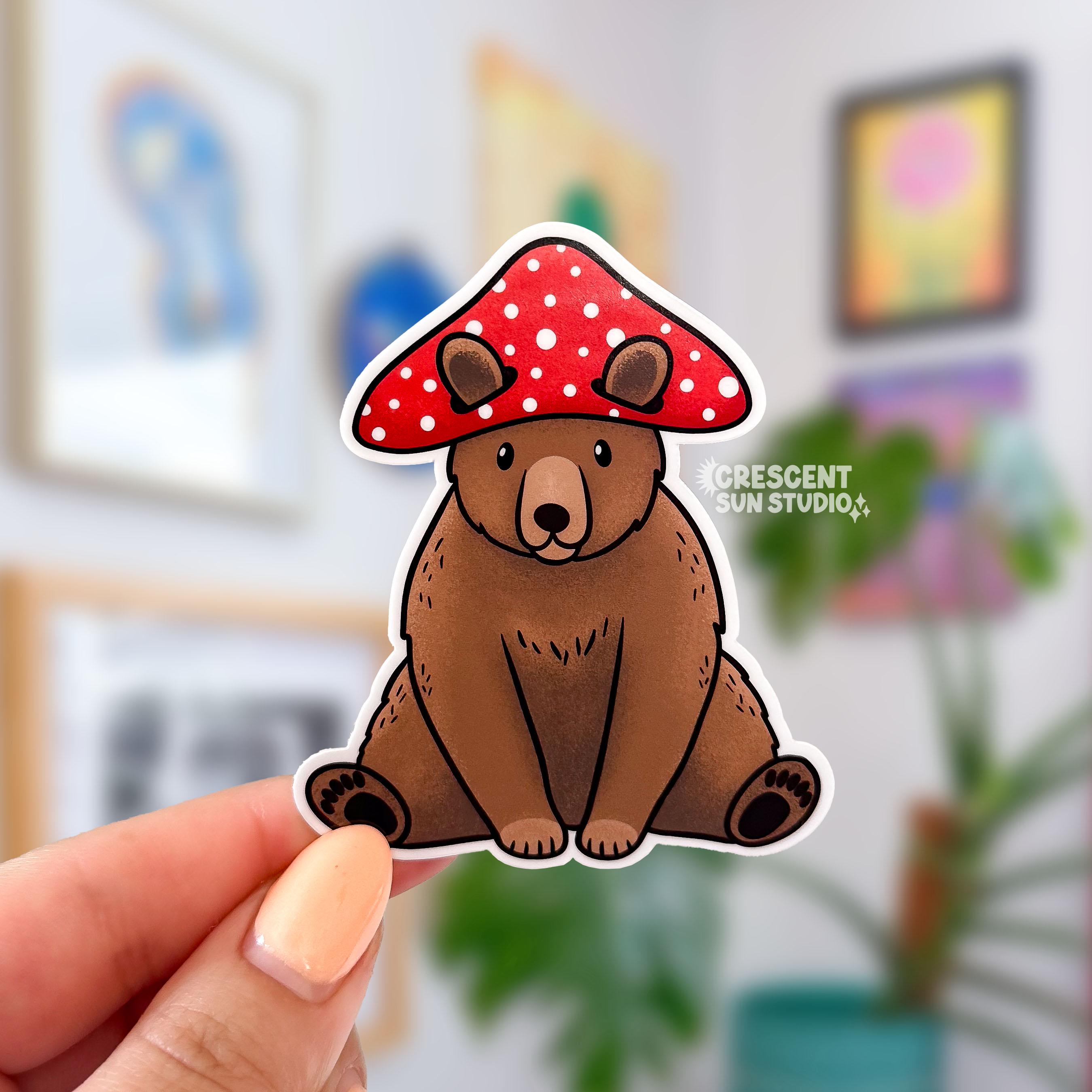 A die cut sticker of a brown bear sitting down and wearing a red and white mushroom hat.