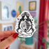 This is a die cut black and white sticker of a nature goddess sitting with her legs crossed and cradling a bee hive.