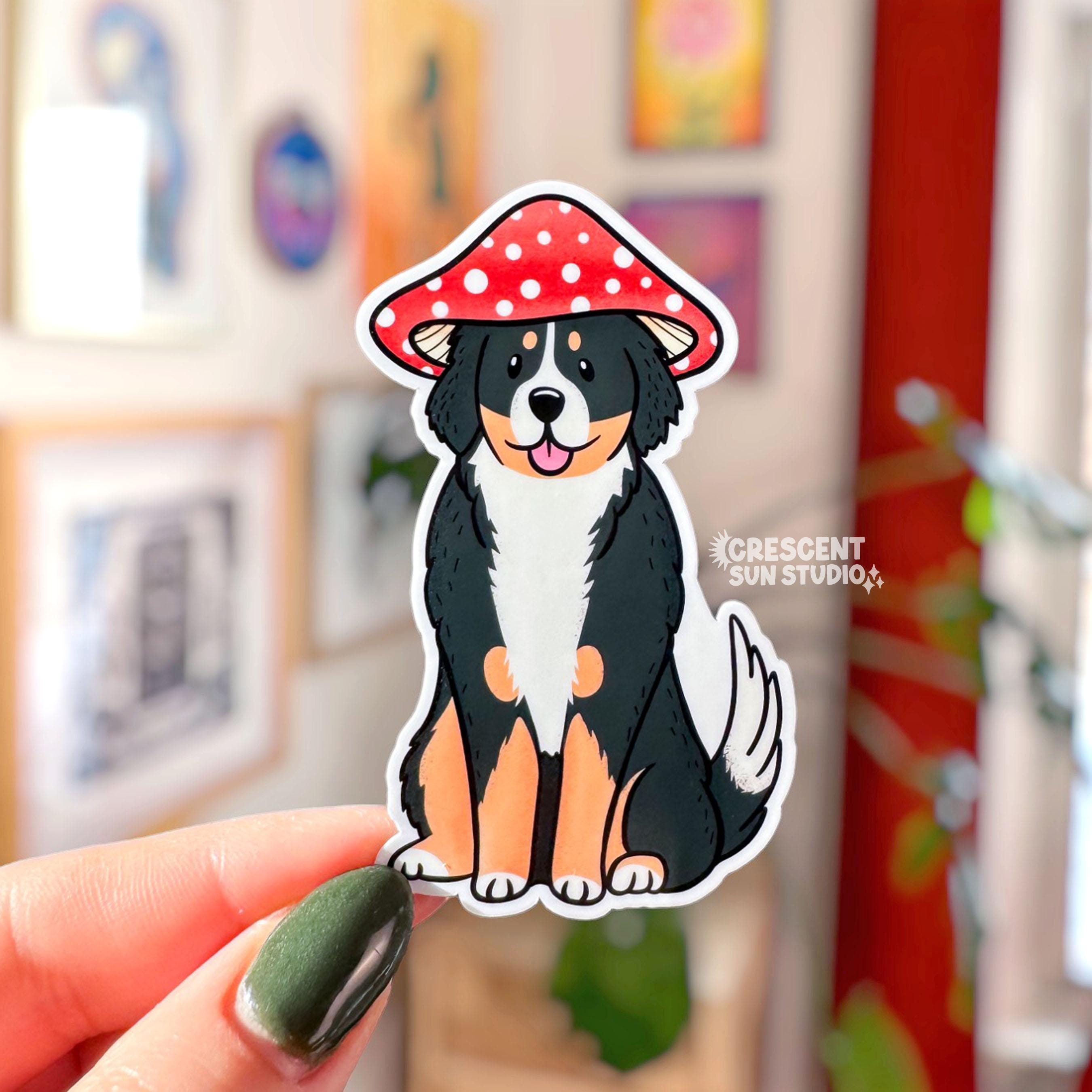 A die cut sticker of a fluffy Bernese mountain dog wearing a red and white mushroom cap for a hat. The dog has a silly expression and his tongue is sticking out.