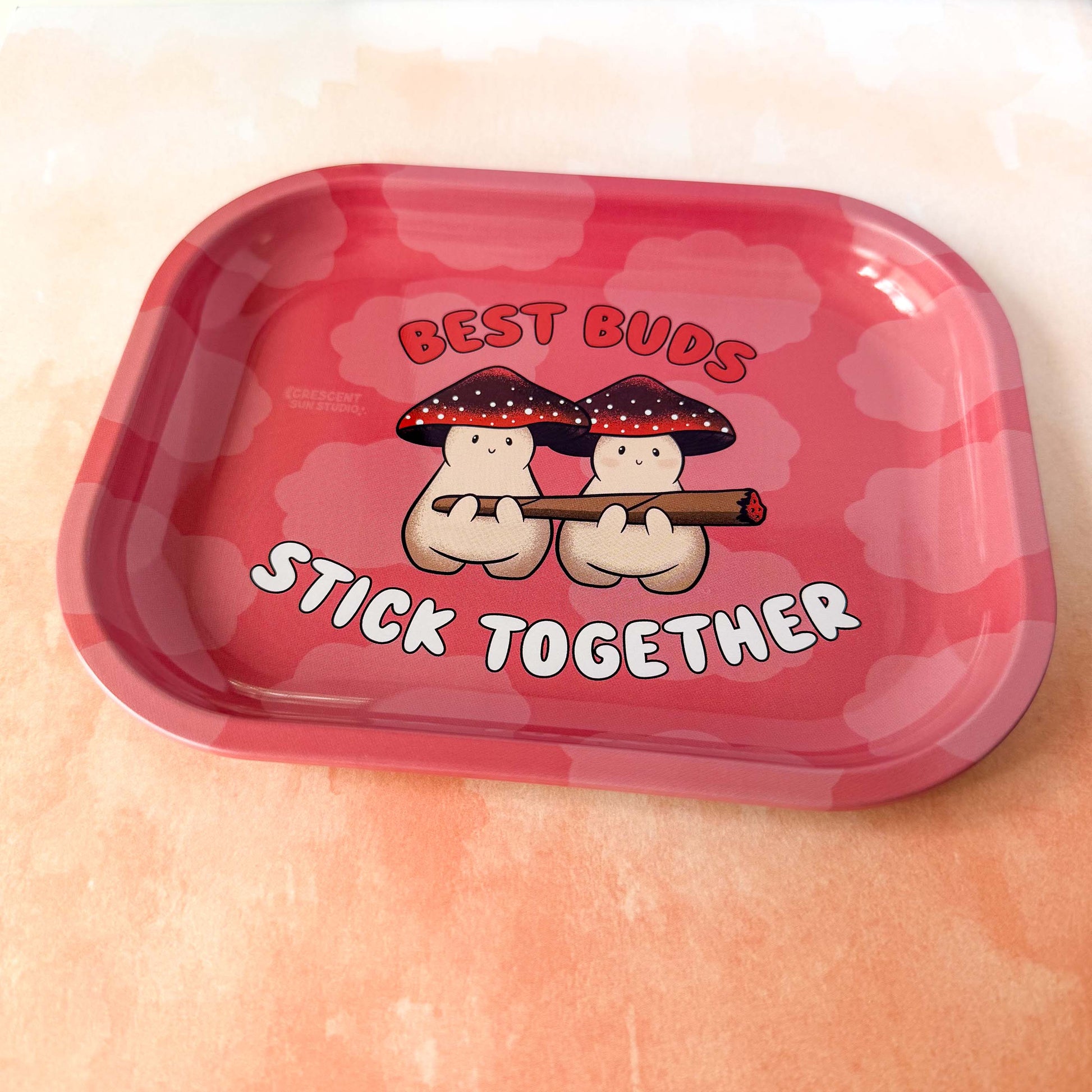 A small red rolling tray with lighter red clouds features two mushroom characters holding a large joint. Both mushroom characters have red and black mushroom caps. The text around them reads "Best Buds Stick Together" in red and white.