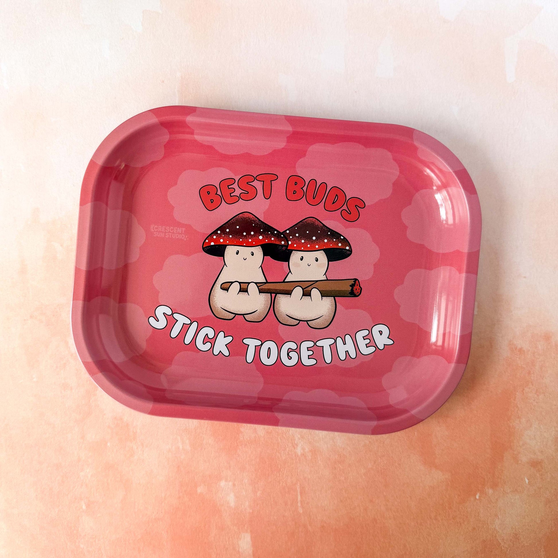 A small red rolling tray with lighter red clouds features two mushroom characters holding a large joint. Both mushroom characters have red and black mushroom caps. The text around them reads "Best Buds Stick Together" in red and white.