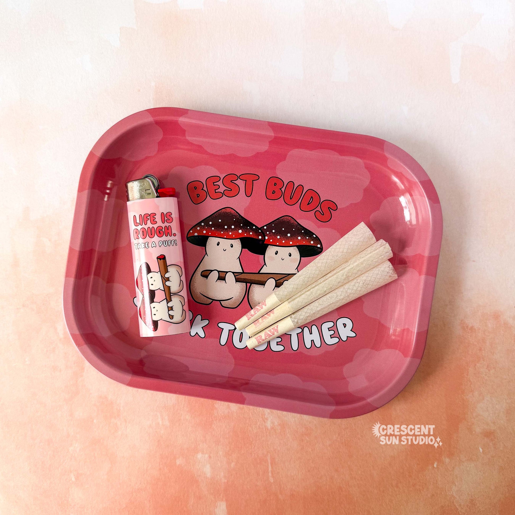 A small red rolling tray with lighter red clouds features two mushroom characters holding a large joint. Both mushroom characters have red and black mushroom caps. The text around them reads "best buds stick together."