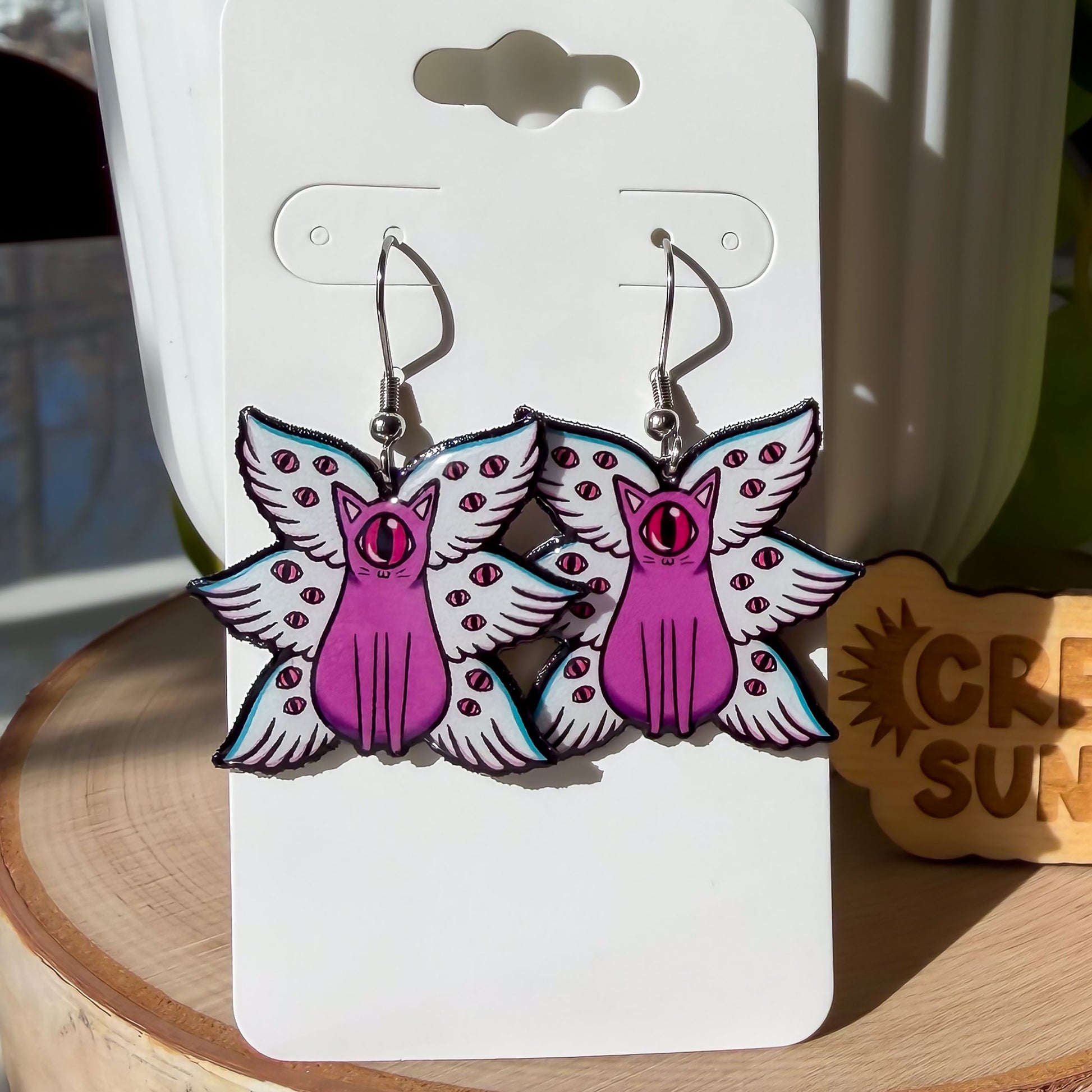 A pair of our biblically accurate cat angels earrings feature purple cats, each with one large pink eye at the center of its stoic face. Each cat has three pairs of light blue angel wings covered with watchful pink eyes.
