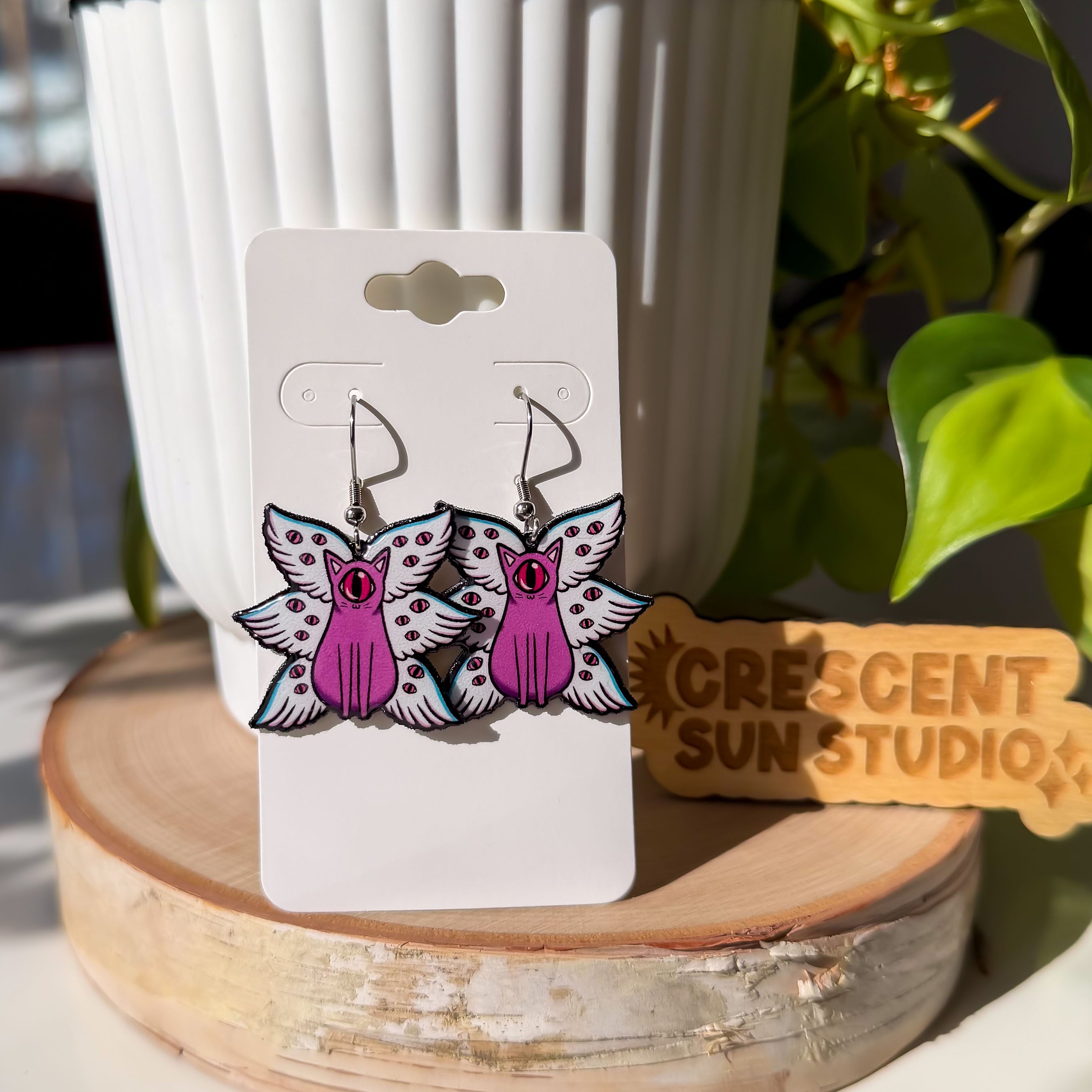 A pair of our biblically accurate cat angels earrings feature purple cats, each with one large pink eye at the center of its stoic face. Each cat has three pairs of light blue angel wings covered with watchful pink eyes.

The earrings hang on a white display card and there is a wooden sign that says "crescent sun studio" in the background.