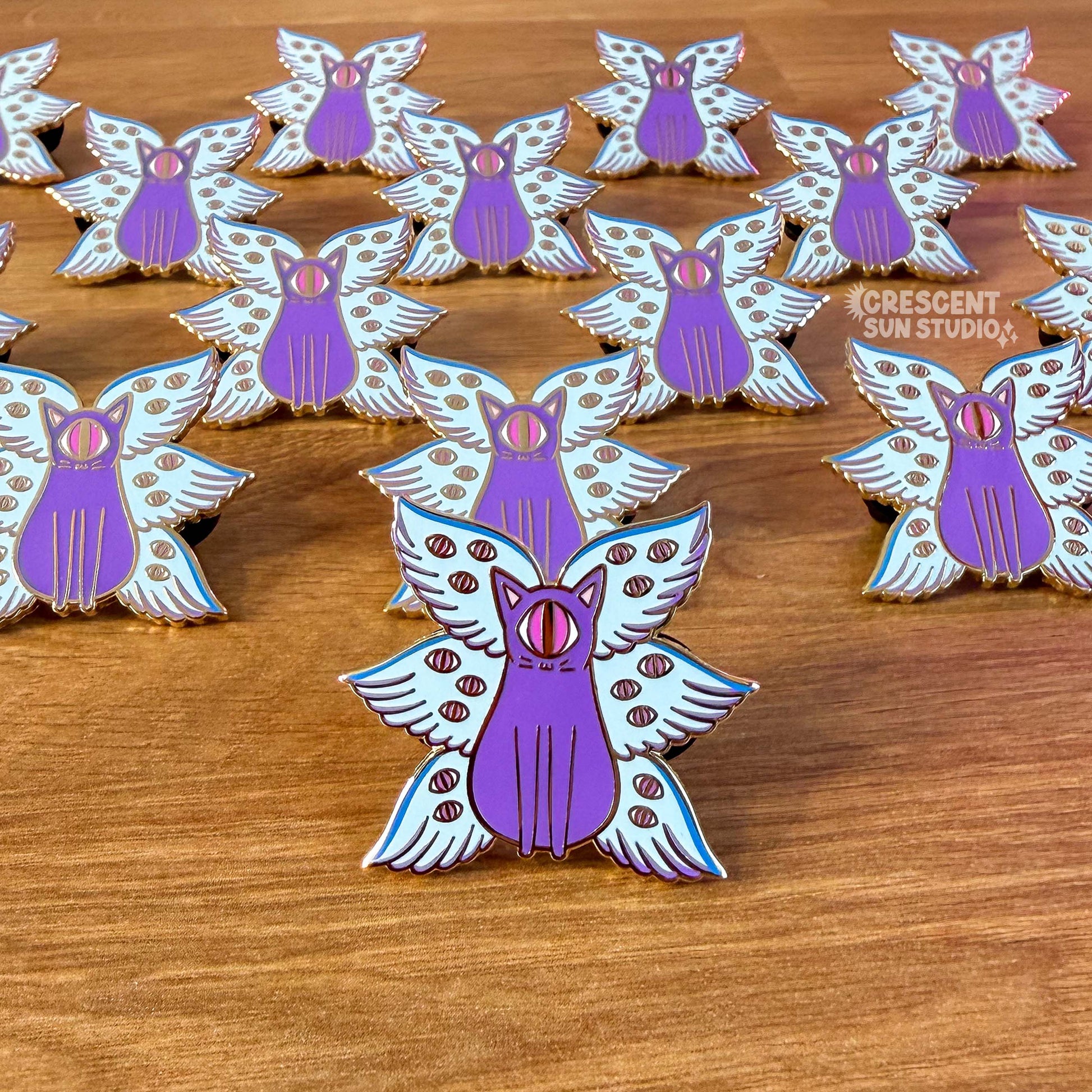 A group of enamel pins that have a purple cat with one large pink eye at the center of its face stoically observes you. The cat has three pairs of light-colored angel wings, each wing covered with watchful pink eyes. 
