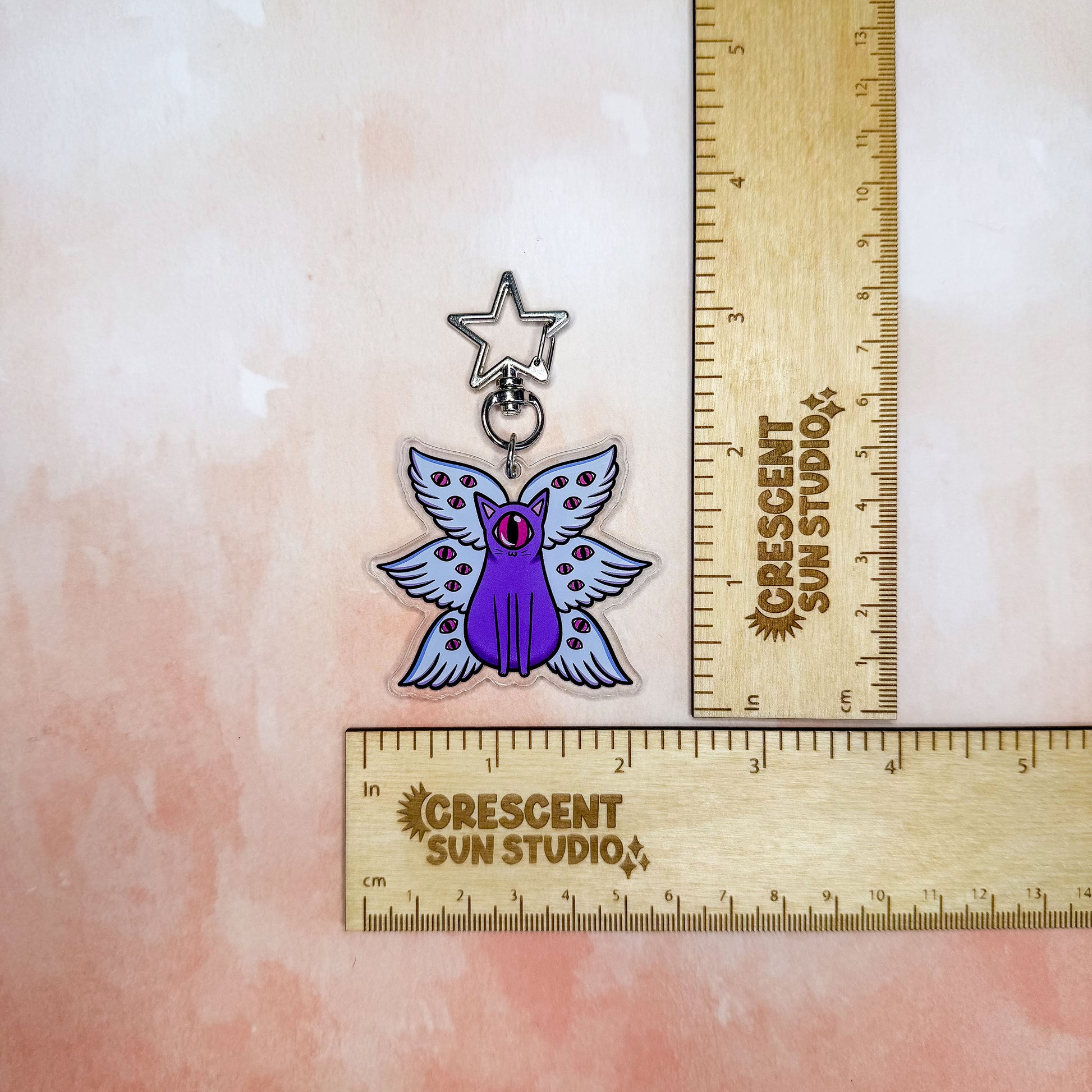 A purple cat with one large pink eye at the center of its face stoically observes you. The cat has three pairs of light-colored angel wings, each wing covered with watchful pink eyes. 

The key chain connects to your favorite things via a star shaped clasp.

This image shows the keychain next to two rulers, showing its size of 2.25 in x 2.25 in