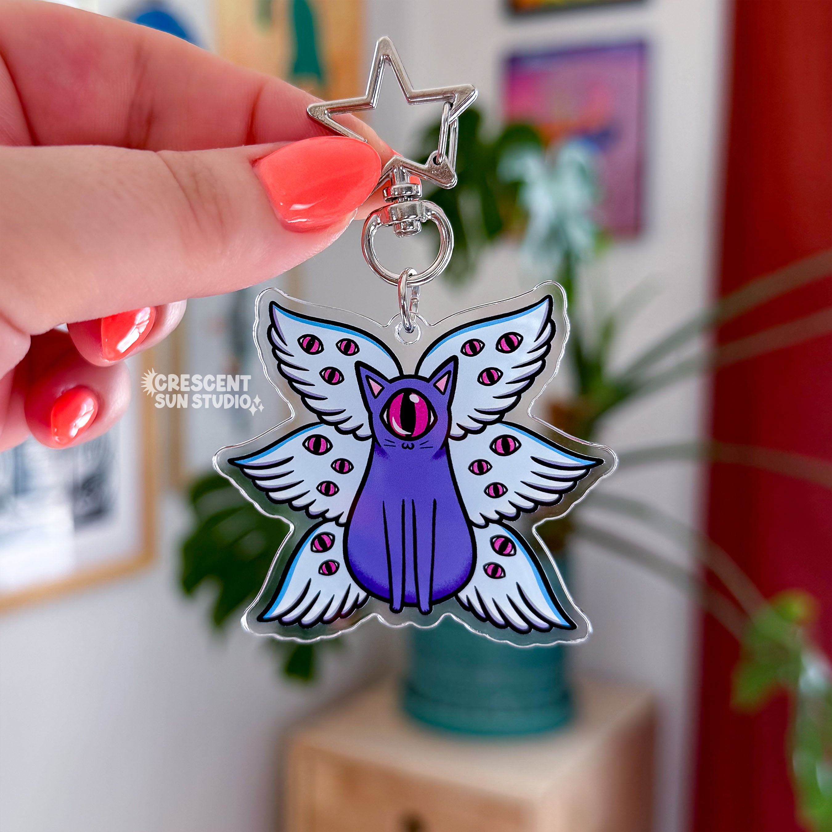 A purple cat with one large pink eye at the center of its face stoically observes you. The cat has three pairs of light-colored angel wings, each wing covered with watchful pink eyes. 

The key chain connects to your favorite things via a star shaped clasp.
