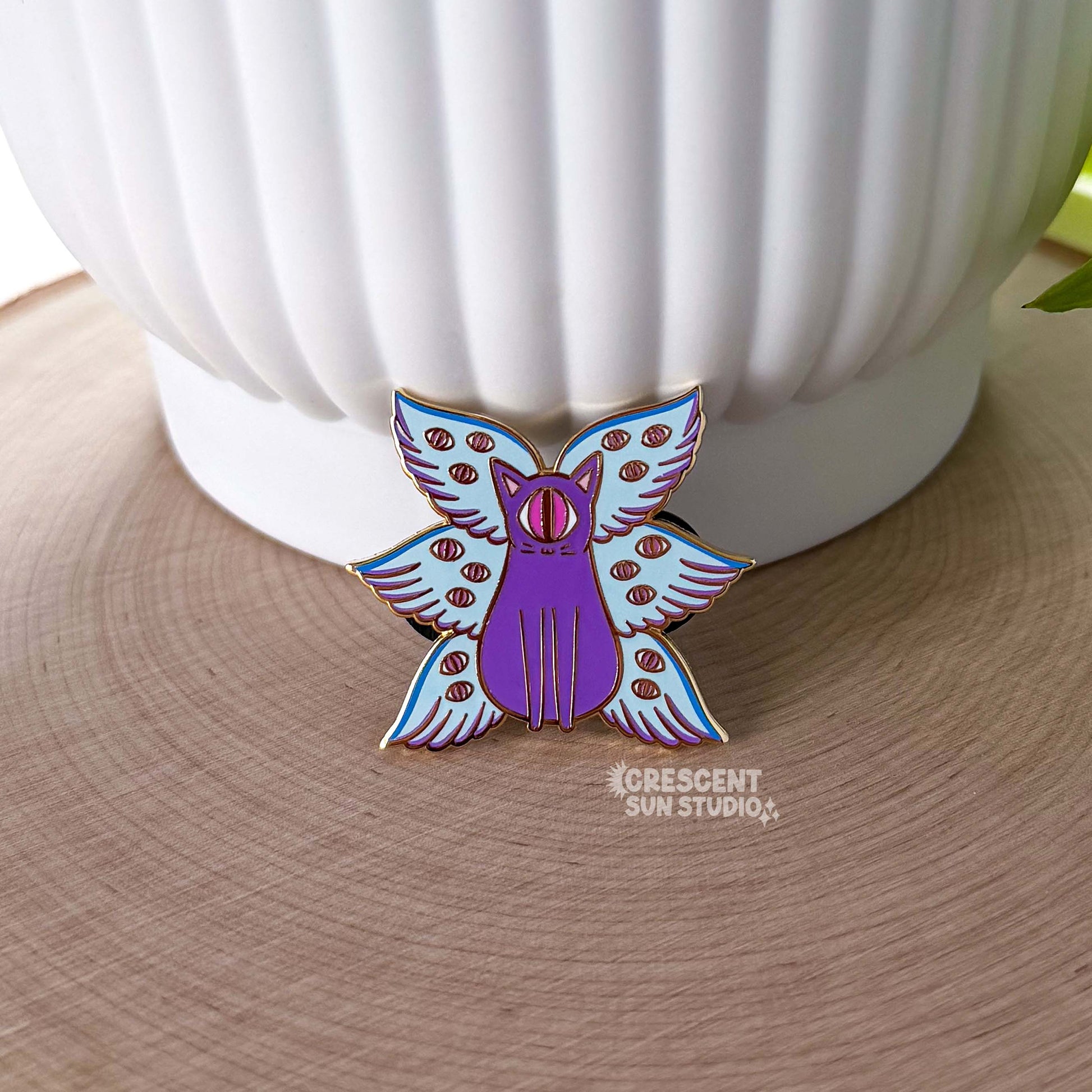 A pin leans against a white pot. The pin is of a purple cat with one large pink eye at the center of its face stoically observes you. The cat has three pairs of light-colored angel wings, each wing covered with watchful pink eyes. 
