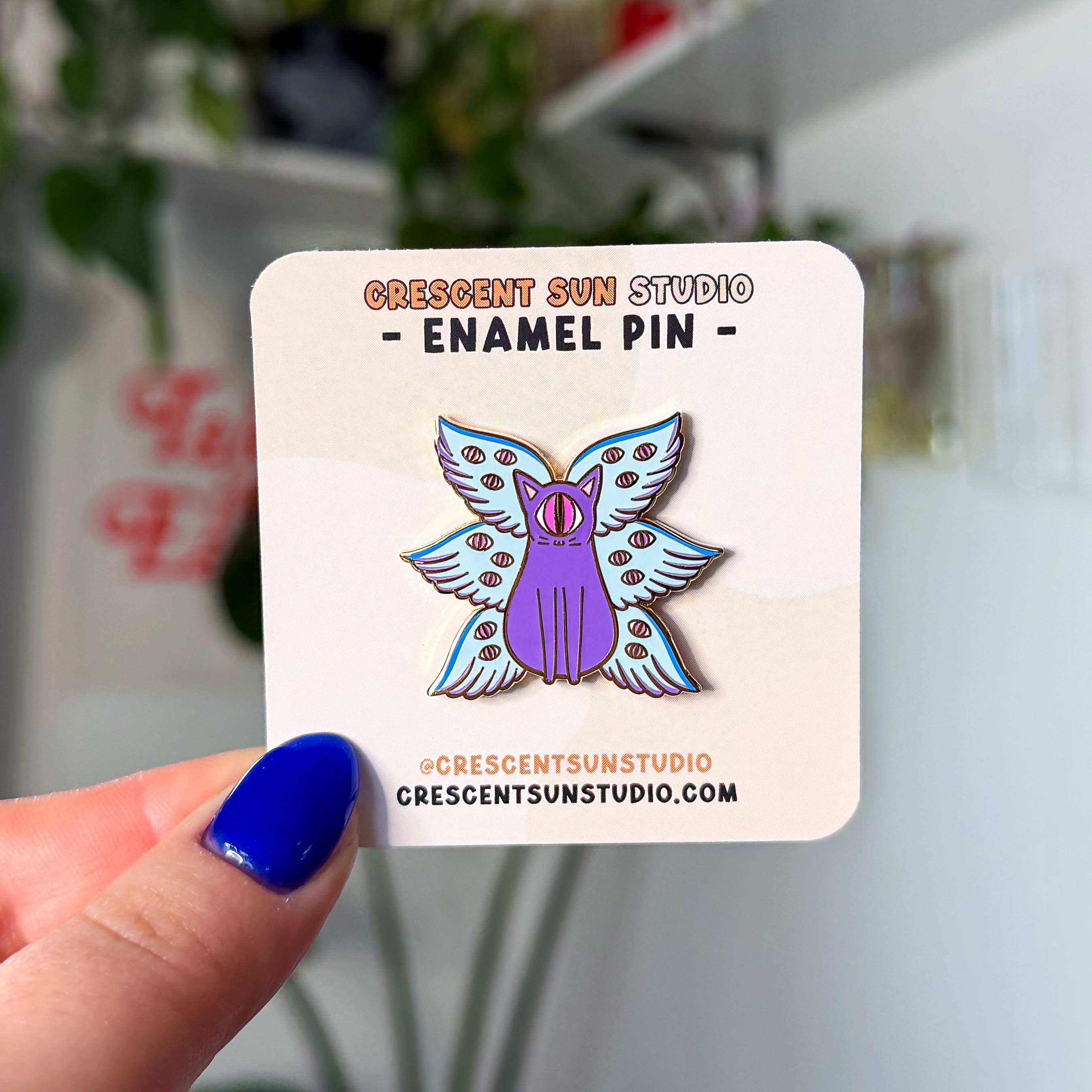 A pin of a purple cat with one large pink eye at the center of its face stoically observes you. The cat has three pairs of light-colored angel wings, each wing covered with watchful pink eyes. 
