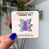 A pin of a purple cat with one large pink eye at the center of its face stoically observes you. The cat has three pairs of light-colored angel wings, each wing covered with watchful pink eyes. 
