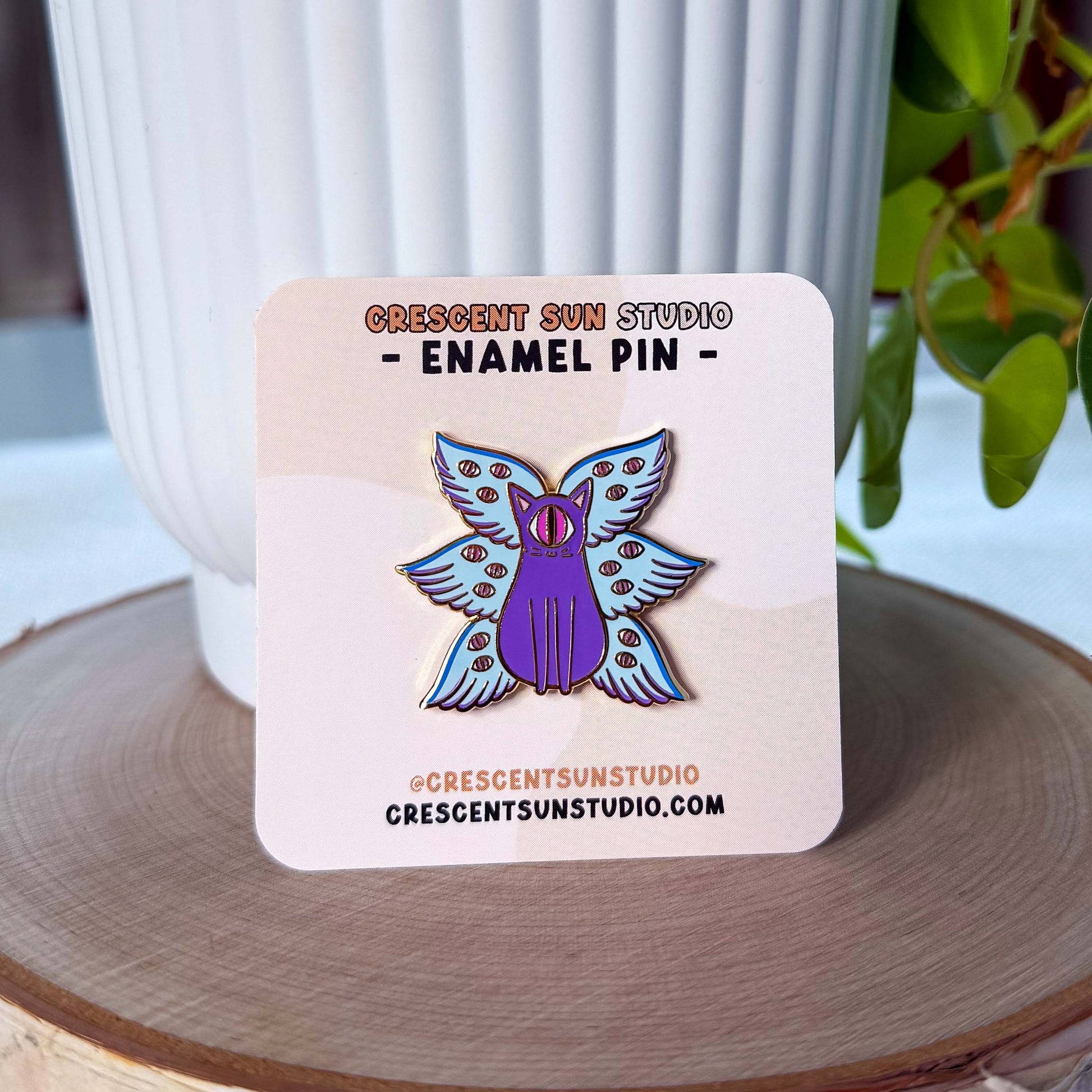 An enamel pin of a purple cat with one large pink eye at the center of its face stoically observes you. The cat has three pairs of light-colored angel wings, each wing covered with watchful pink eyes. 
