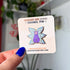 A small enamel pin of a purple cat with one large pink eye at the center of its face stoically observes you. The cat has three pairs of light-colored angel wings, each wing covered with watchful pink eyes. 
