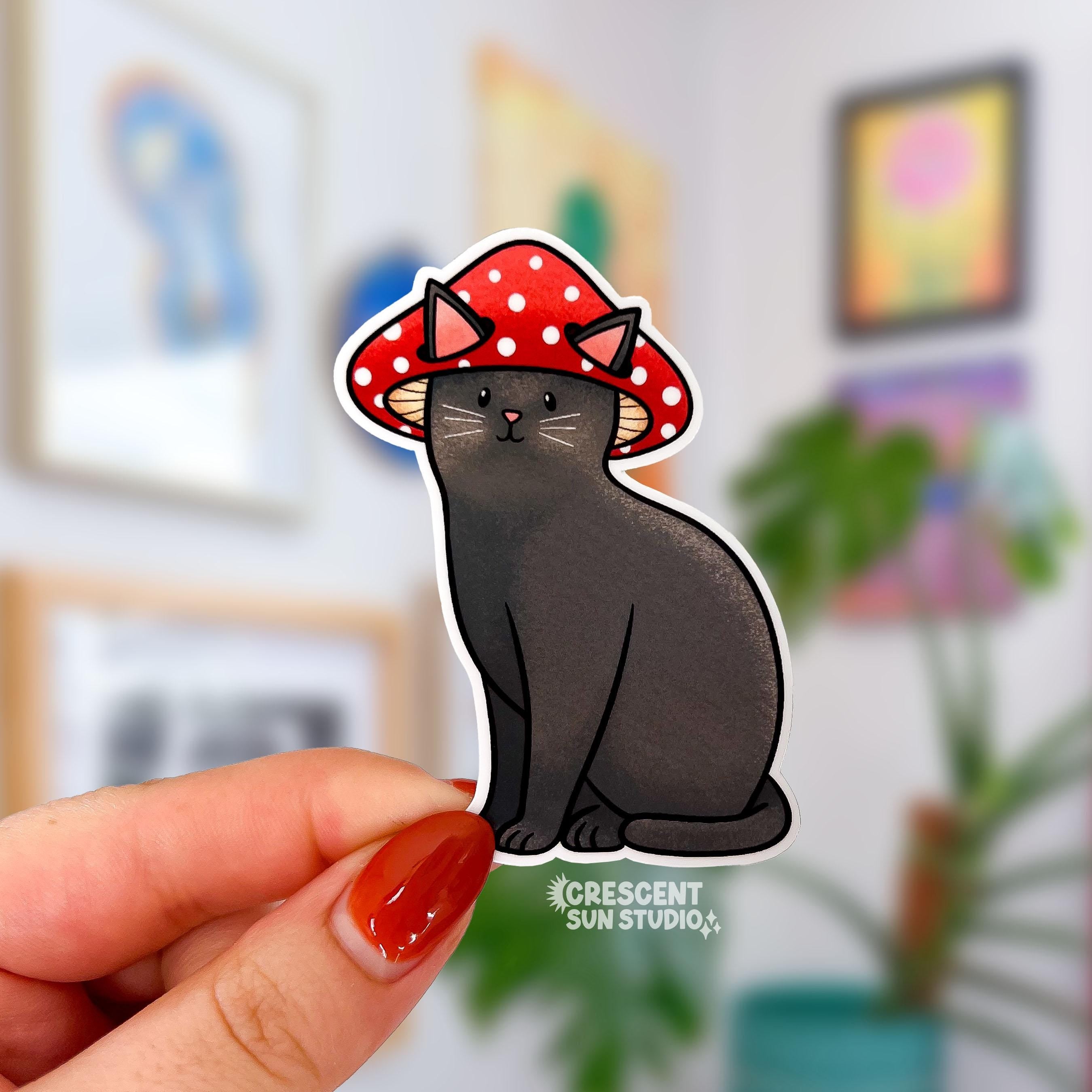 A sticker in the shape of a sitting cat is held close to the camera. The cat is black with white whiskers and has a contented look on his face. He is wearing a red mushroom cap with white spots and his large pink earspoke through holes in the cap.