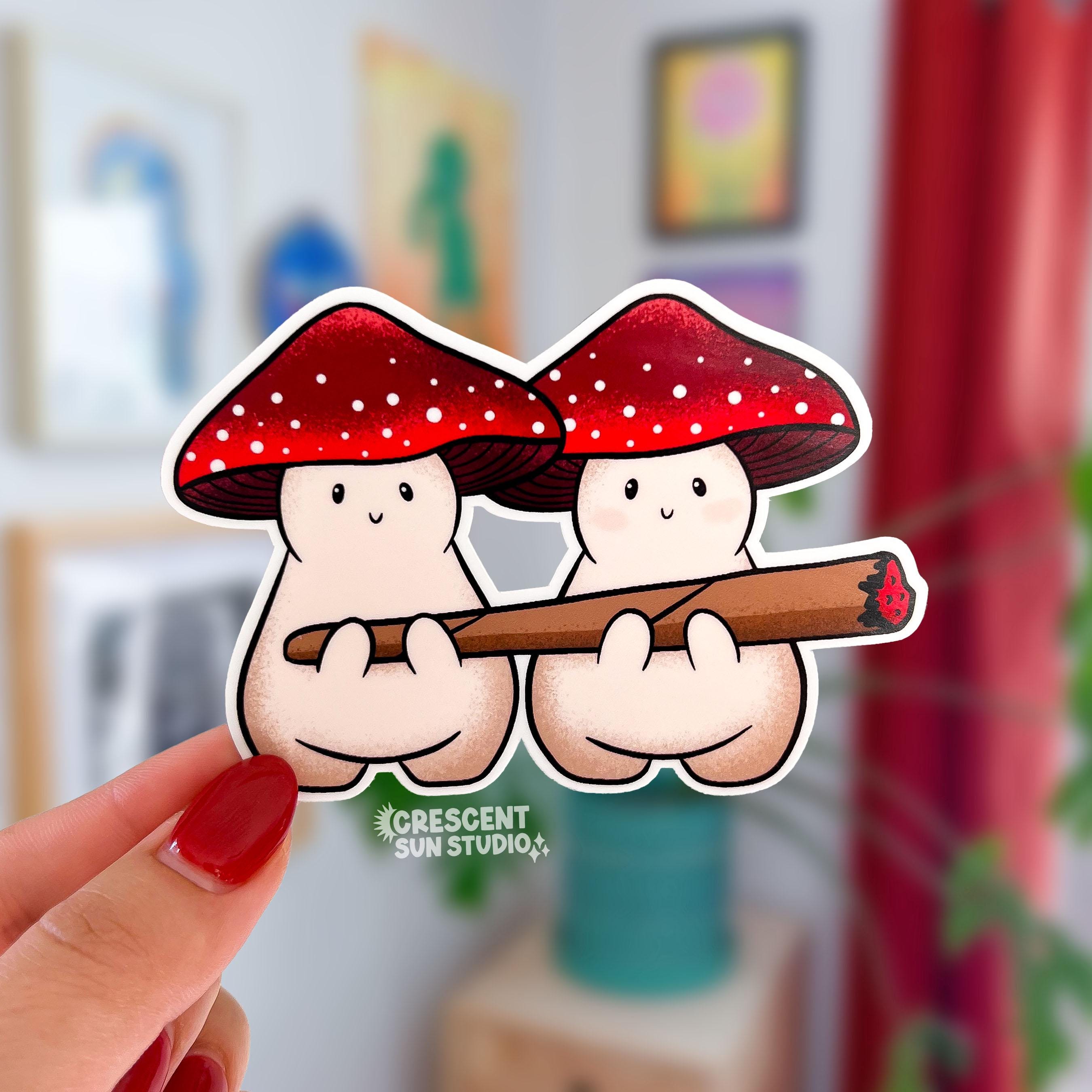 Die cut sticker of two plump beige mushroom characters with red and white spotted caps, sit side by side sharing a lit blunt.