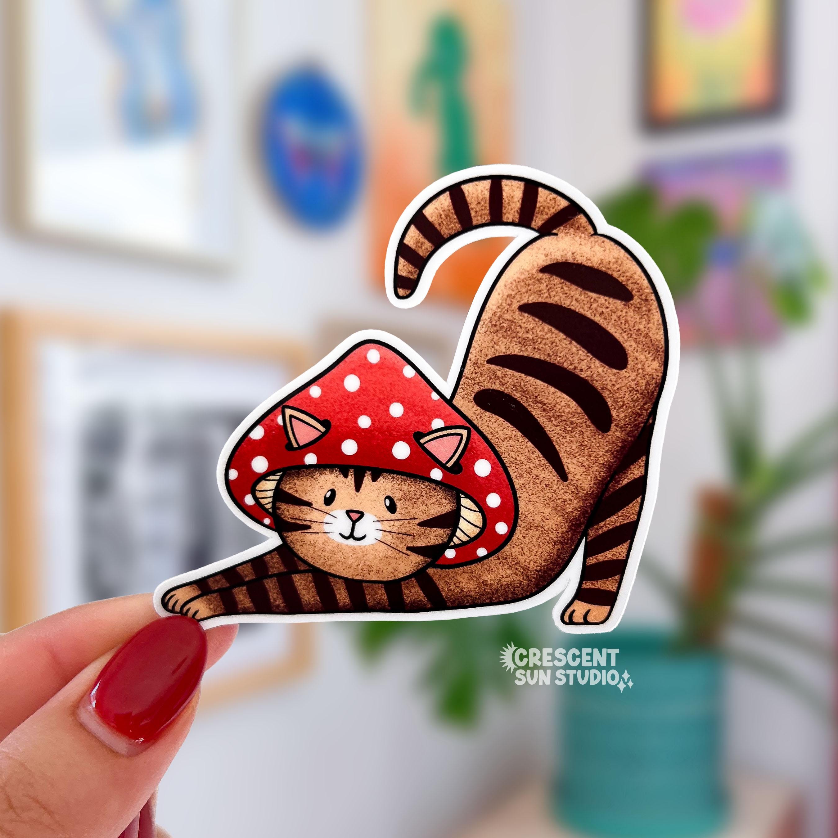 A sticker in the shape of a cat wearing a mushroom hat is held close to the camera. The cat is a brown tabby (medium brown with darker brown stripes) and has a sweet expression on his face. His red mushroom hat with white spots has spots for his ears to poke through. The cat is in a stretching pose with his tail curling up behind him.