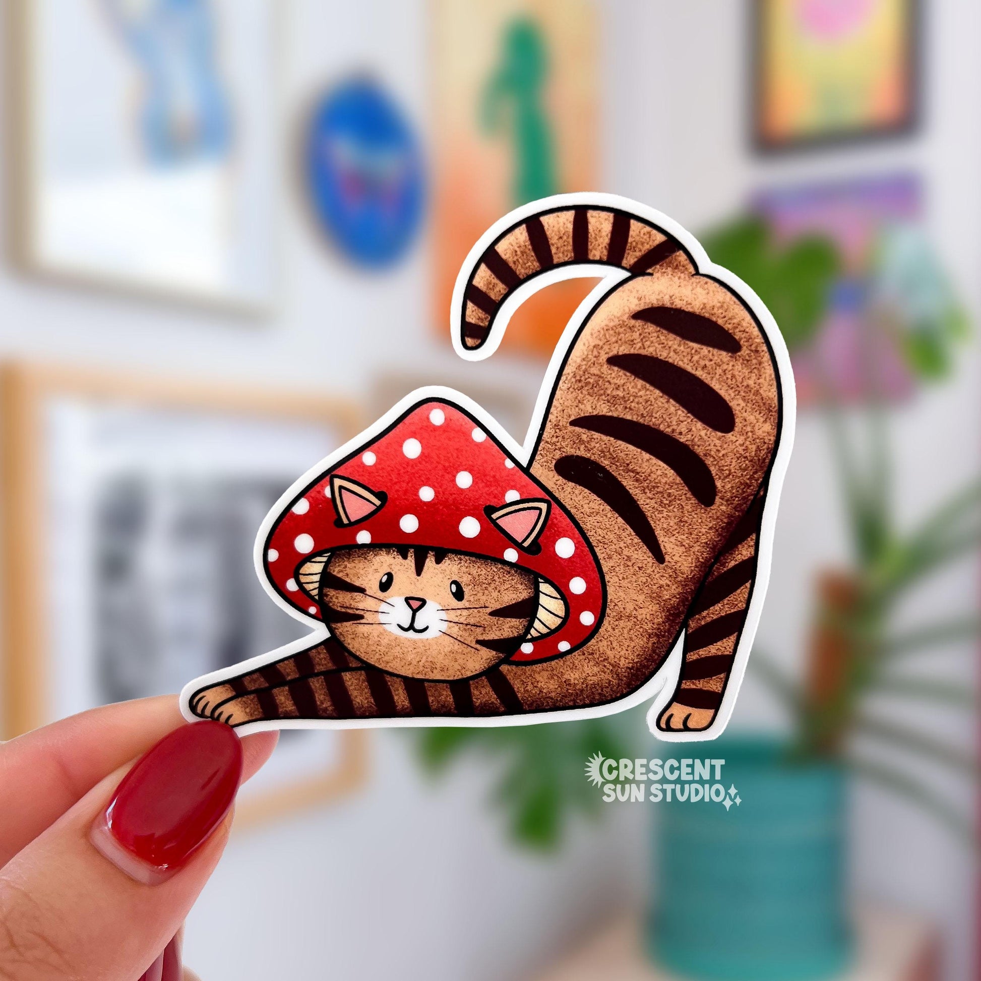 A sticker in the shape of a cat wearing a mushroom hat is held close to the camera. The cat is a brown tabby (medium brown with darker brown stripes) and has a sweet expression on his face. His red mushroom hat with white spots has spots for his ears to poke through. The cat is in a stretching pose with his tail curling up behind him.