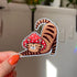 A sticker in the shape of a cat wearing a mushroom hat is held close to the camera. The cat is a brown tabby (medium brown with darker brown stripes) and has a sweet expression on his face. His red mushroom hat with white spots has spots for his ears to poke through. The cat is in a stretching pose with his tail curling up behind him.

In this image, a woman's hand holds the kitty sticker up to the camera.
