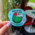 A blue die cut sticker of a cactus cup with a straw, red flower, and cactus juice surrounded by the quotes, "drink cactus juice," "nothing's quenchier," "it's the quenchiest," and "it'll quench ya!"