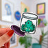 Die-cut water-resistant sticker of a smiling cannabis nug sitting inside an open glass jar with a purple lid.