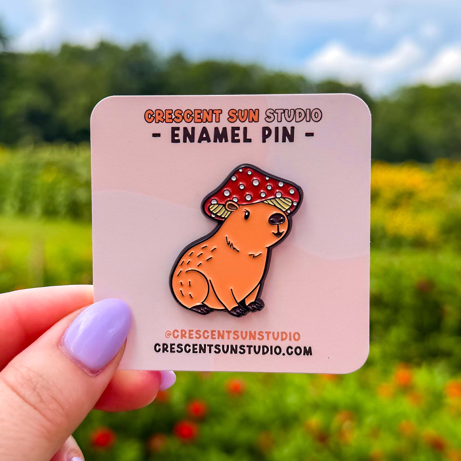 A small pin in the shape of a sitting capybara wearing a mushroom cap on his head. The capybara is brown and has a happy expression on his face. His cap is red with white spots. The pin is on a light pink display card and held up to the camera.
