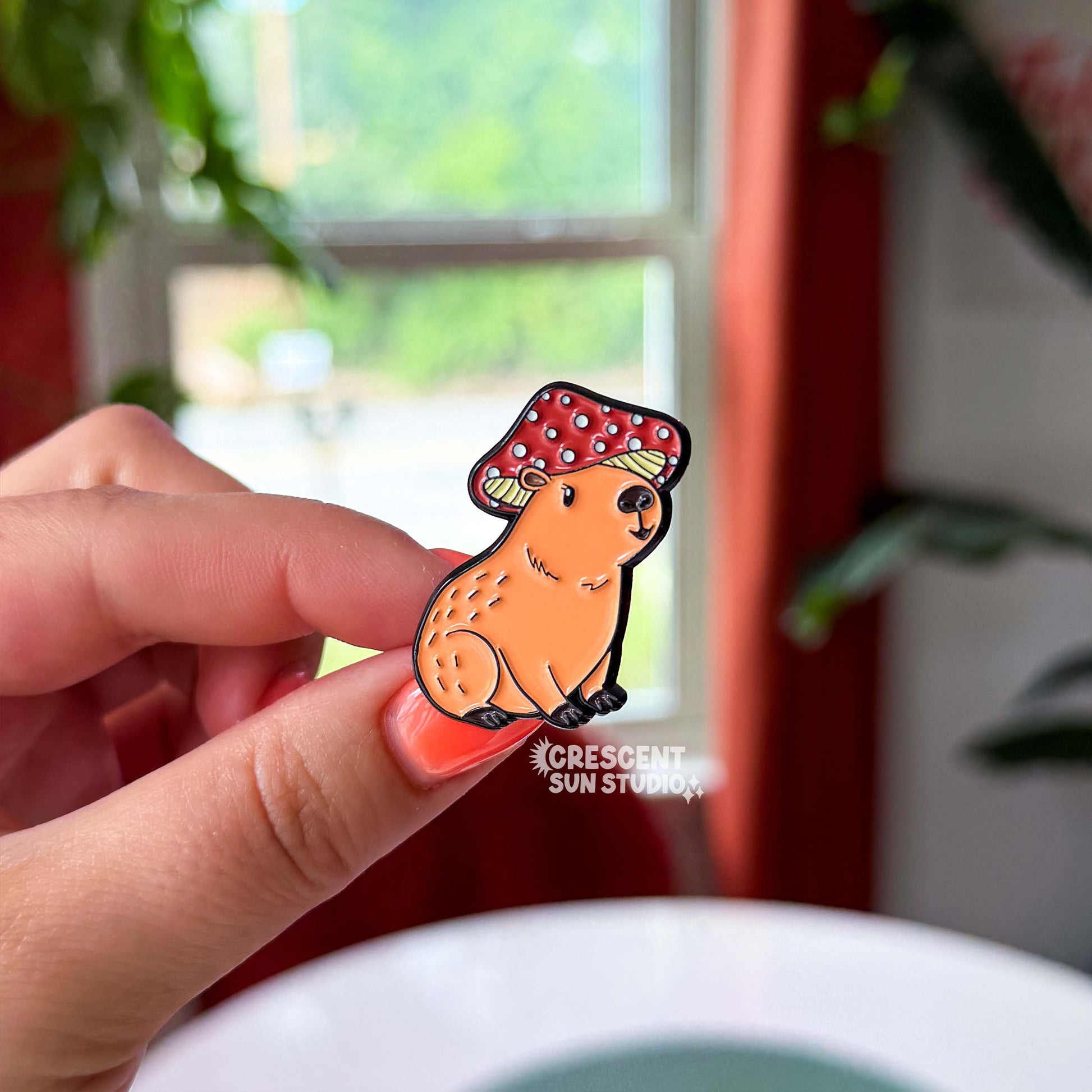 A small pin in the shape of a sitting capybara wearing a mushroom cap on his head. The capybara is brown and has a happy expression on his face. His cap is red with white spots.