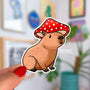 A sticker in the shape of a sitting capybara wearing a mushroom cap on his head. The capybara is brown and has a happy expression on his face. His cap is red with white spots.