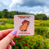 A small pin in the shape of a sitting capybara wearing a mushroom cap on his head. The capybara is brown and has a happy expression on his face. His cap is red with white spots. The pin is on a light pink display card and held up to the camera.