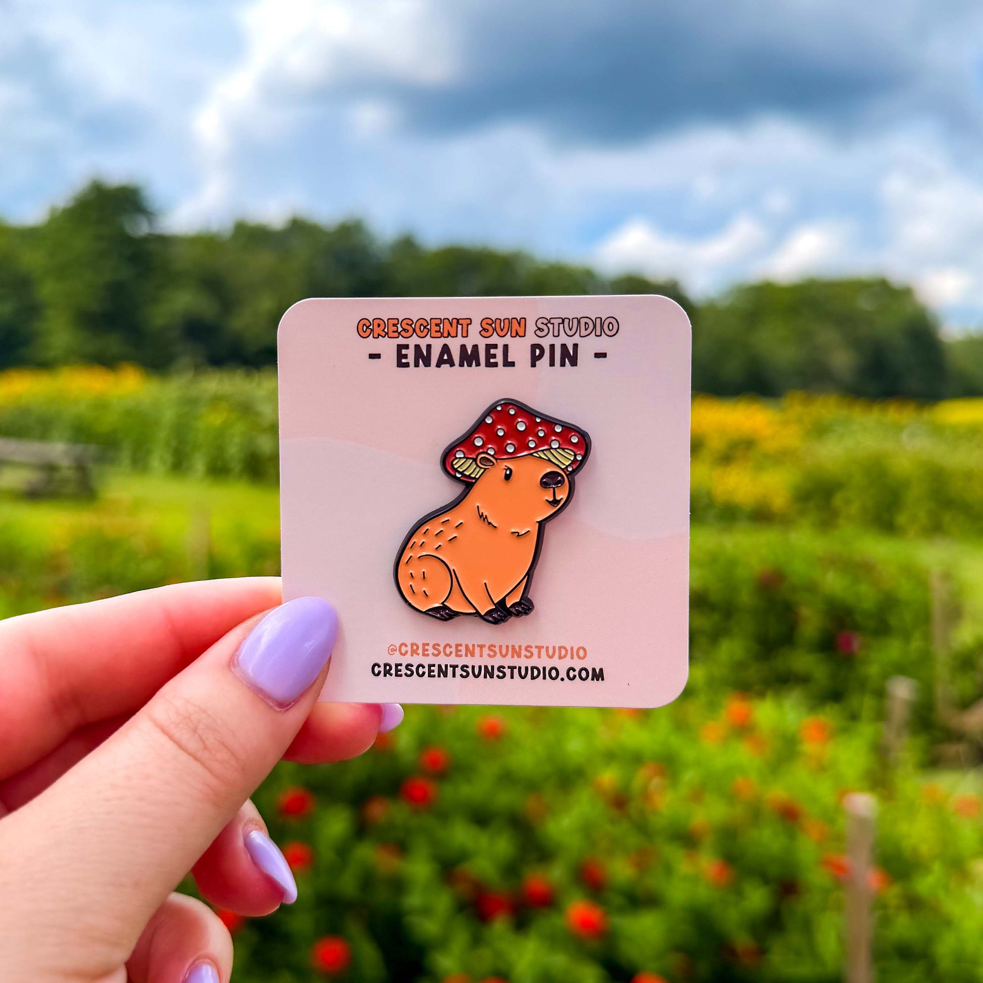 A small pin in the shape of a sitting capybara wearing a mushroom cap on his head. The capybara is brown and has a happy expression on his face. His cap is red with white spots. The pin is on a light pink display card and held up to the camera.
