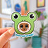 This sticker depicts the face of a brown cat with tears welling up in her eyes. She has pink cheeks and long whiskers. She is wearing a bright green frog head-shaped hoodie, with giant black frog eyes. The sticker is in the shape of the frog head. Underneath the design is blue text that says "certified crybaby."