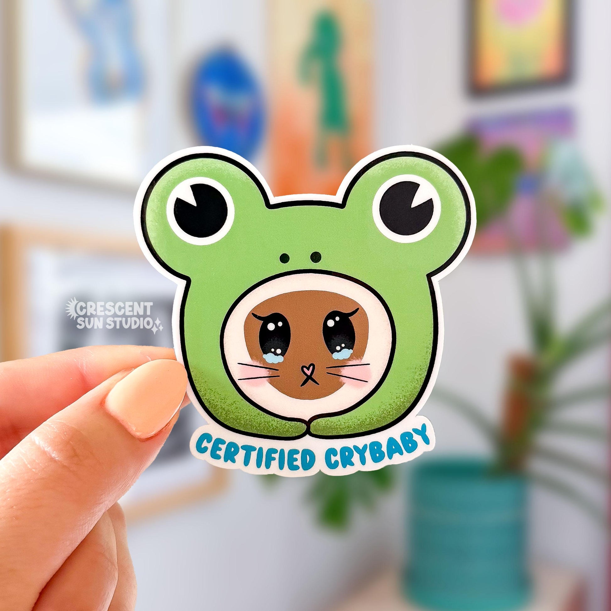 This sticker depicts the face of a brown cat with tears welling up in her eyes. She has pink cheeks and long whiskers. She is wearing a bright green frog head-shaped hoodie, with giant black frog eyes. The sticker is in the shape of the frog head. Underneath the design is blue text that says "certified crybaby."