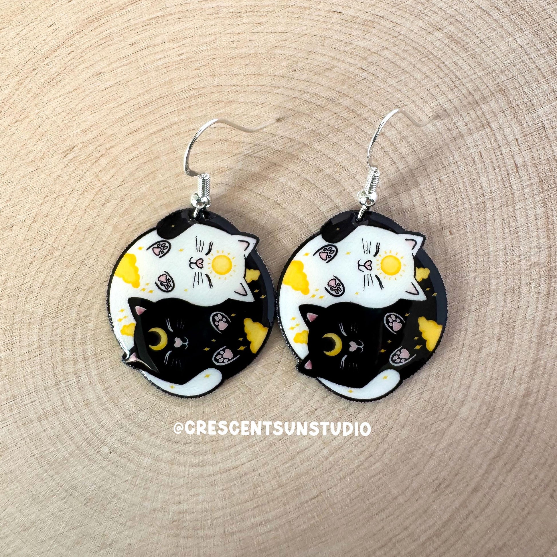This image depicts a pair of round dangle earrings. On each earring is a black cat and a white cat curled up in a ying yang symbol. The black cat has a yellow crescent moon between her eyes and the white cat has a sun symbol between her eyes. Both smiling cats have two paws pointed outward, so you can see their pink toe pads.