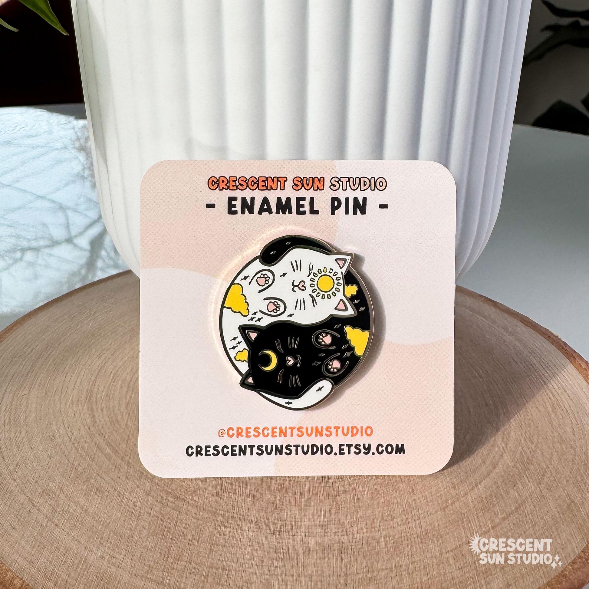 A round enamel pin depicting a black cat and a white cat curled up in a yin yang symbol. The black cat has a yellow crescent moon between her eyes and the white cat has a sun symbol between her eyes. Both smiling cats have two paws pointed outward, so you can see their pink toe pads.

In this image, the pin is displayed on a light colored card, which is propped up against a white plant pot.