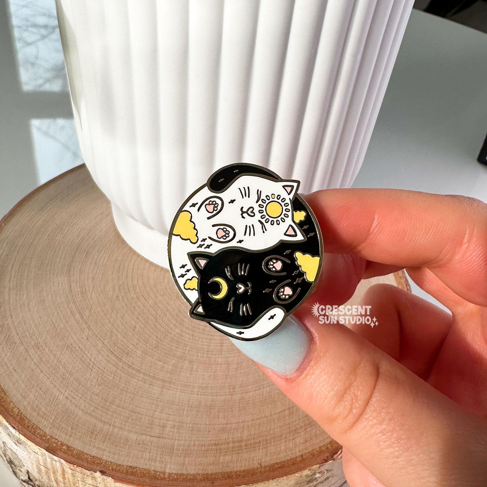 A round enamel pin depicting a black cat and a white cat curled up in a yin yang symbol. The black cat has a yellow crescent moon between her eyes and the white cat has a sun symbol between her eyes. Both smiling cats have two paws pointed outward, so you can see their pink toe pads.

In this image, the pin is held up to the camera by a woman's hand. 