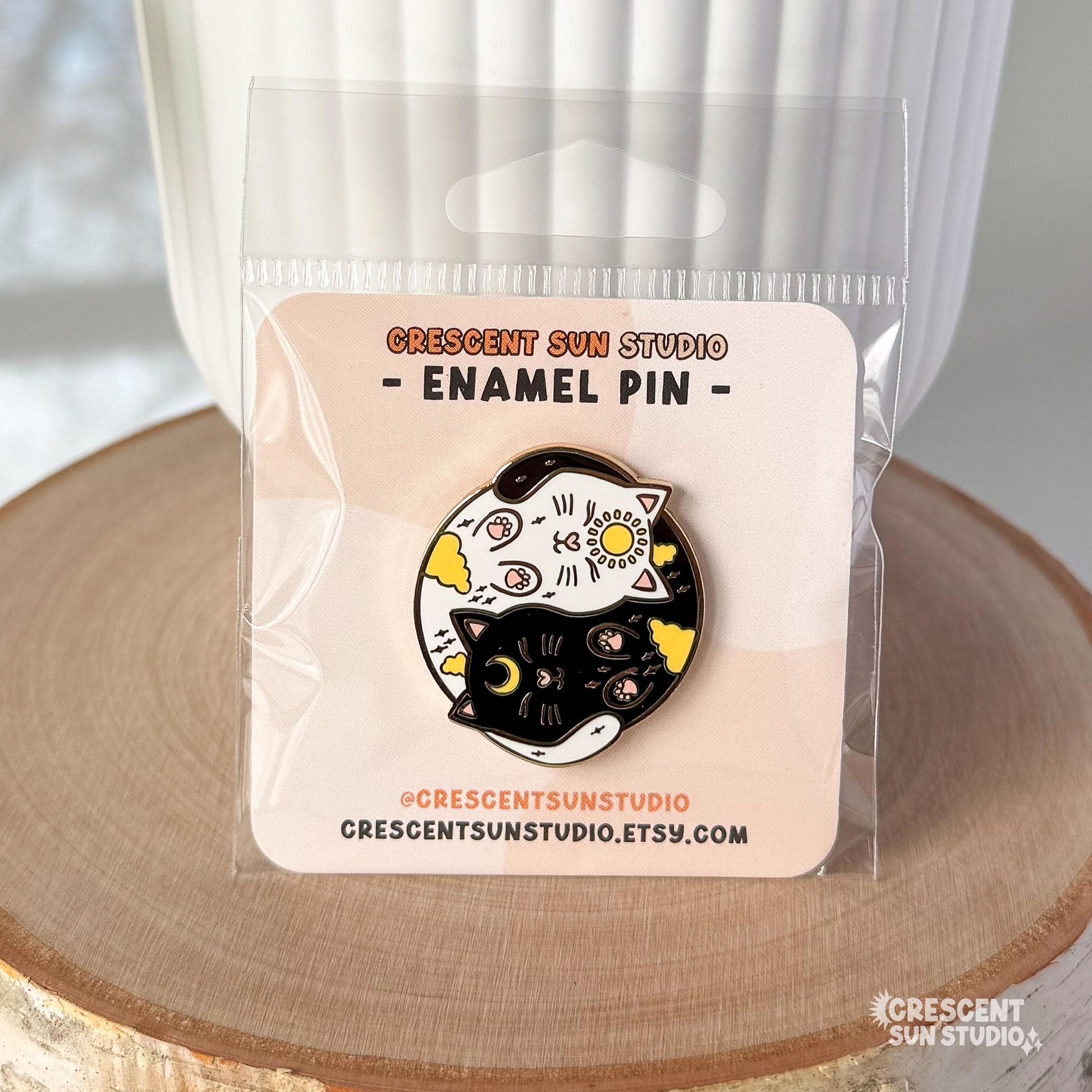 A round enamel pin depicting a black cat and a white cat curled up in a yin yang symbol. The black cat has a yellow crescent moon between her eyes and the white cat has a sun symbol between her eyes. Both smiling cats have two paws pointed outward, so you can see their pink toe pads.

This image shows the clear plastic bag the pin arrives in.