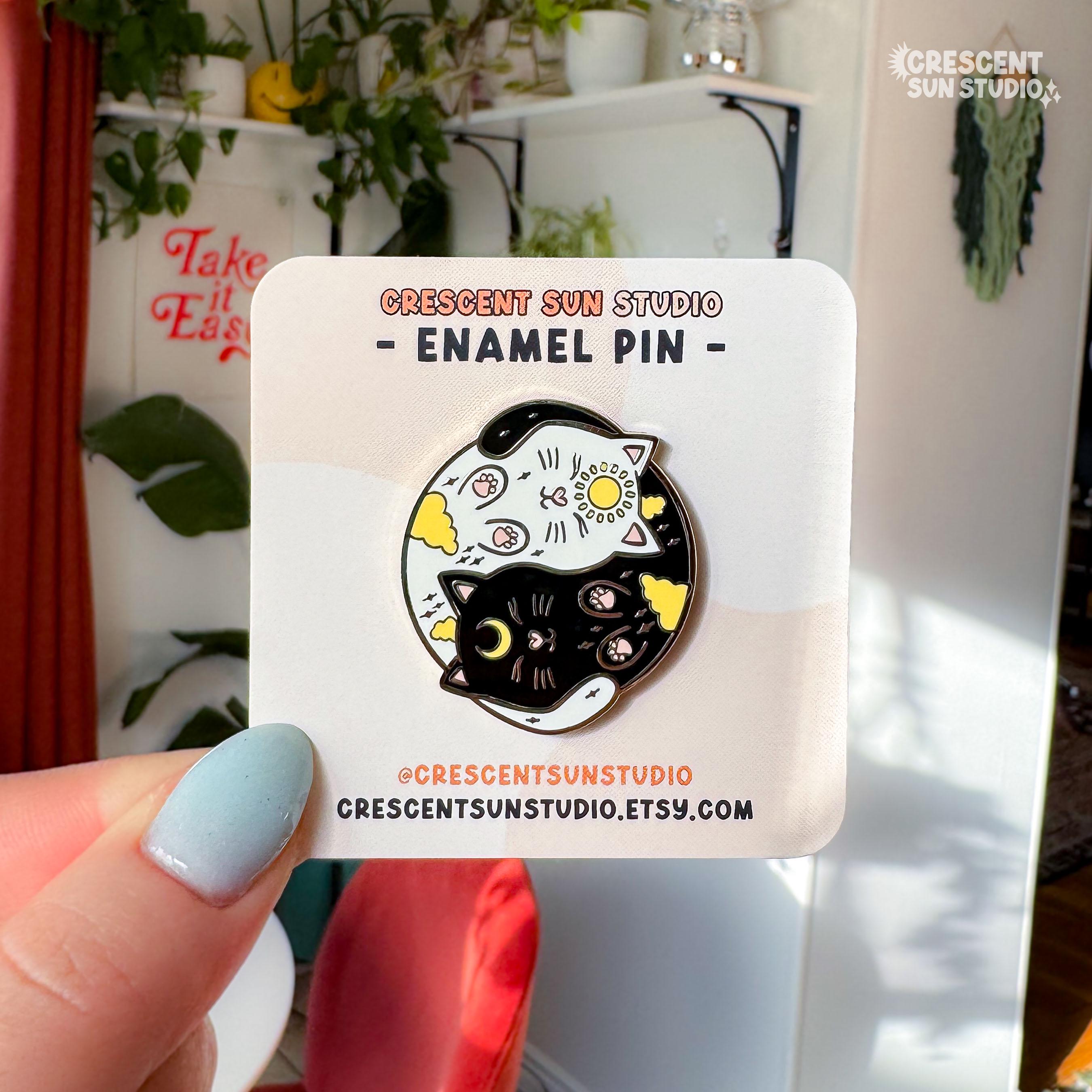 A round enamel pin depicting a black cat and a white cat curled up in a yin yang symbol. The black cat has a yellow crescent moon between her eyes and the white cat has a sun symbol between her eyes. Both smiling cats have two paws pointed outward, so you can see their pink toe pads.

In this image, the pin is displayed on a light colored card and held up to the camera.