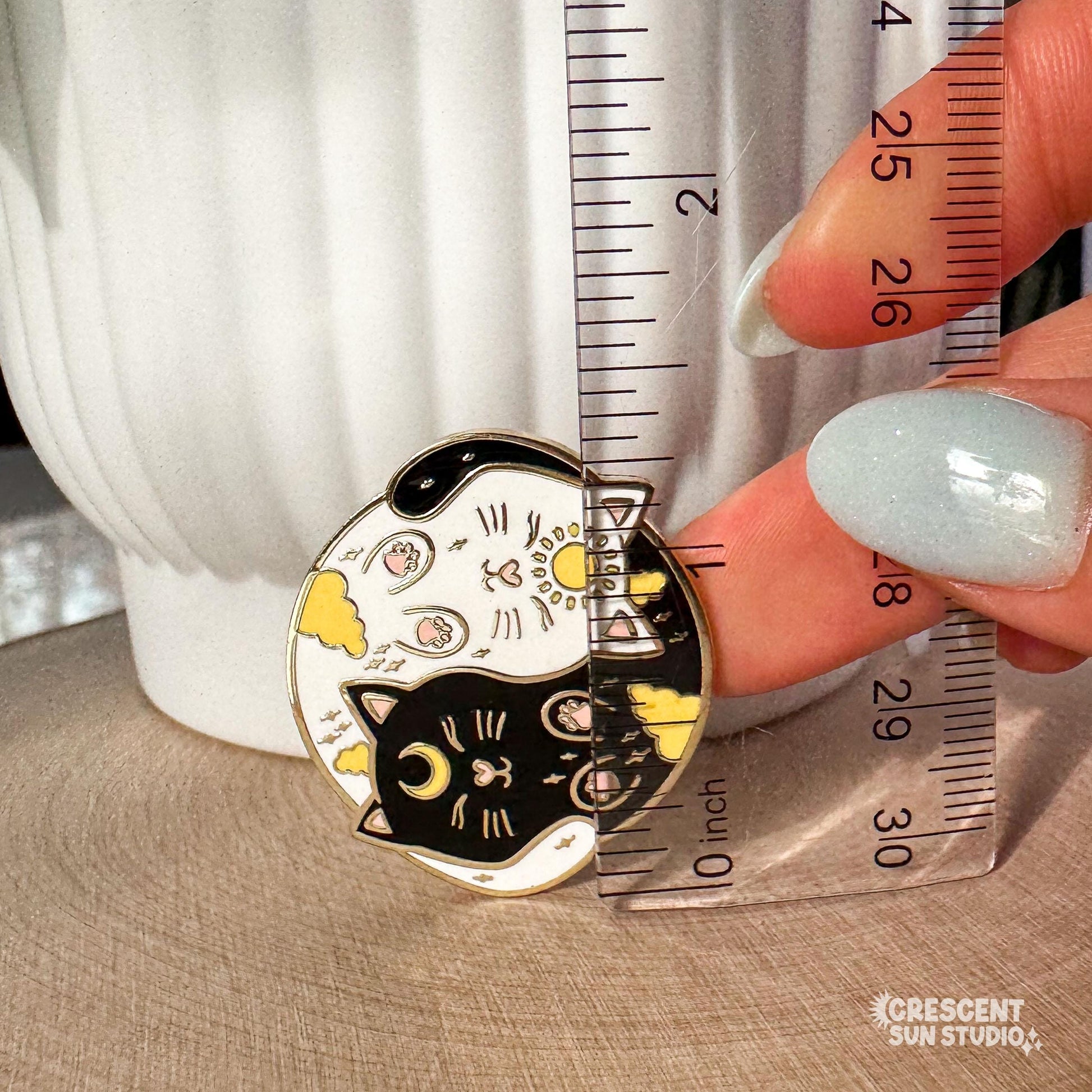 A round enamel pin depicting a black cat and a white cat curled up in a yin yang symbol. The black cat has a yellow crescent moon between her eyes and the white cat has a sun symbol between her eyes. Both smiling cats have two paws pointed outward, so you can see their pink toe pads.

In this image, the pin is next to a clear plastic ruler, showing its size of 1.35 inches tall.