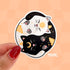A round sticker depicting a black cat and a white cat curled up in a yin yang symbol. The black cat has a yellow crescent moon between her eyes and the white cat has a sun symbol between her eyes. Both smiling cats have two paws pointed outward, so you can see their pink toe pads.