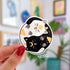 A round sticker depicting a black cat and a white cat curled up in a yin yang symbol. The black cat has a yellow crescent moon between her eyes and the white cat has a sun symbol between her eyes. Both smiling cats have two paws pointed outward, so you can see their pink toe pads.