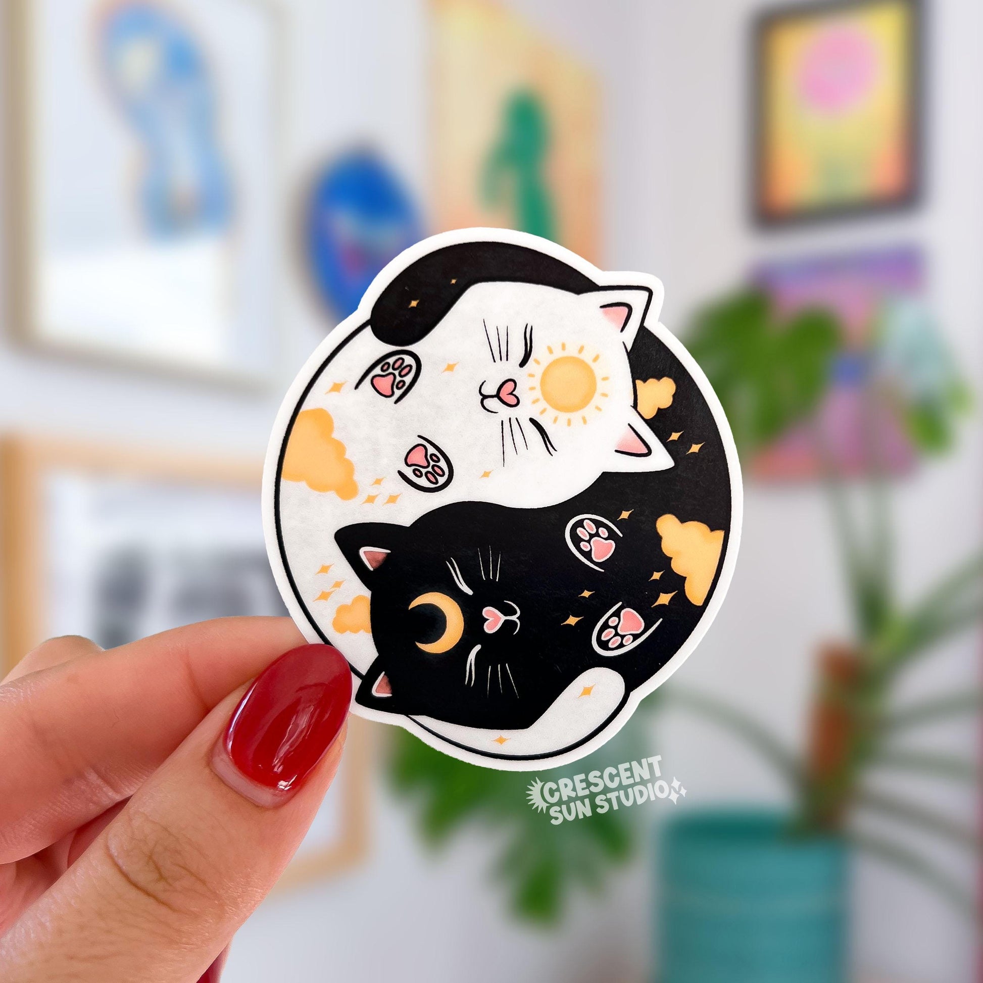 A round sticker depicting a black cat and a white cat curled up in a yin yang symbol. The black cat has a yellow crescent moon between her eyes and the white cat has a sun symbol between her eyes. Both smiling cats have two paws pointed outward, so you can see their pink toe pads.