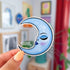 A sticker in the shape of a crescent moon. The sticker is shaded blue and has the profile of a serene and confident woman's face. The face has a thin arched eyebrow, closed eye with deeper blue shadow and lush lashes. She has flushed cheeks and full lips that are glossy purply-blue.
