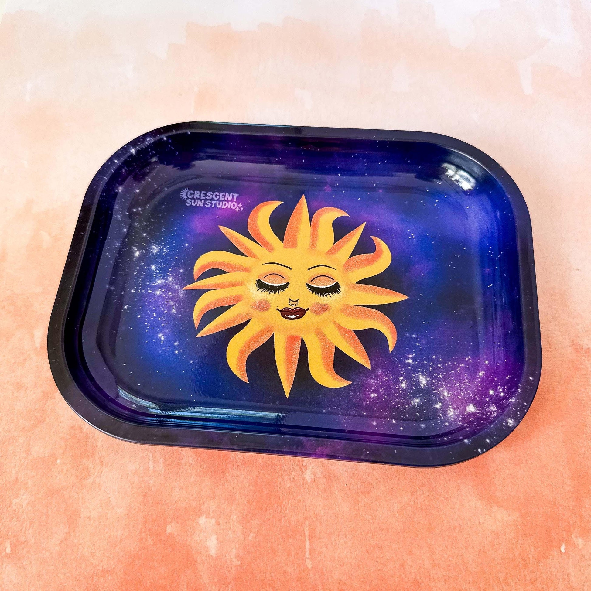 This rectangle tray with raised edges, features a blissed out and elevated glam sun floats in a purple-blue galaxy background. This personified sun has thin eyebrows, closed eyes with full lashes and eyeshadow, a nose ring, blushing cheeks, and glossy full lips.

This image is shot at an angle.