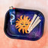 This rectangle tray with raised edges, features a blissed out and elevated glam sun floats in a purple-blue galaxy background. This personified sun has thin eyebrows, closed eyes with full lashes and eyeshadow, a nose ring, blushing cheeks, and glossy full lips.

A lighter with a crescent moon design sits on the tray and three pre-rolled cones sit on the opposite side of the lighter.