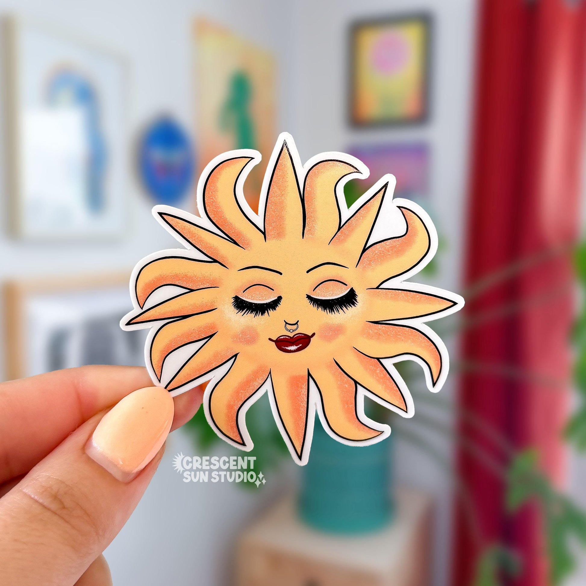 This sticker features a blissed out and glam sun with rays of light dancing around her. This personified sun has thin eyebrows, closed eyes with full lashes and eyeshadow, a nose ring, blushing cheeks, and glossy full lips.