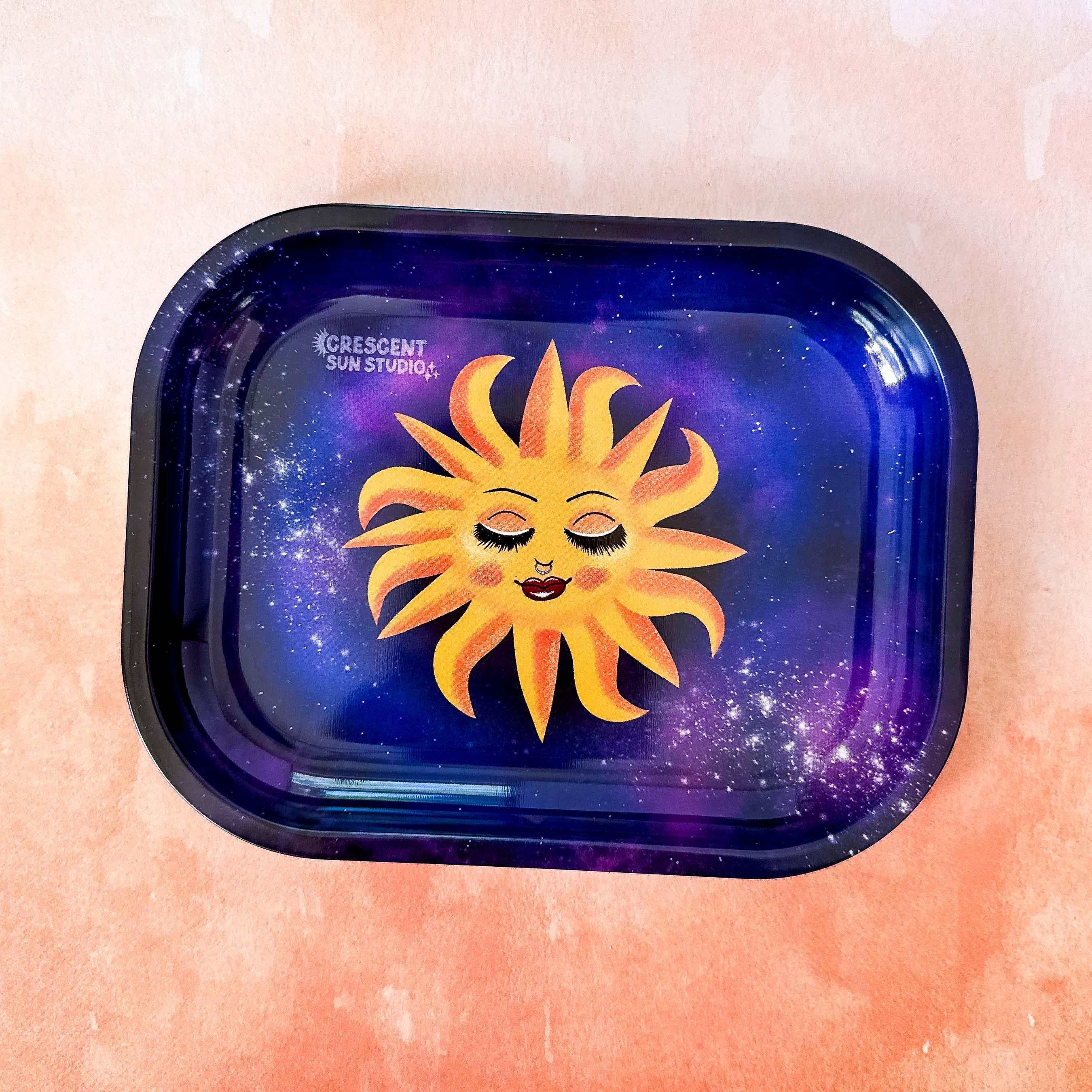 This rectangle tray with raised edges, features a blissed out and elevated glam sun floats in a purple-blue galaxy background. This personified sun has thin eyebrows, closed eyes with full lashes and eyeshadow, a nose ring, blushing cheeks, and glossy full lips.