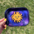 This rectangle tray with raised edges, features a blissed out and elevated glam sun floats in a purple-blue galaxy background. This personified sun has thin eyebrows, closed eyes with full lashes and eyeshadow, blushing cheeks, and glossy full lips.

A hand with yellow nails is holding the tray up to the camera against a green textured background.